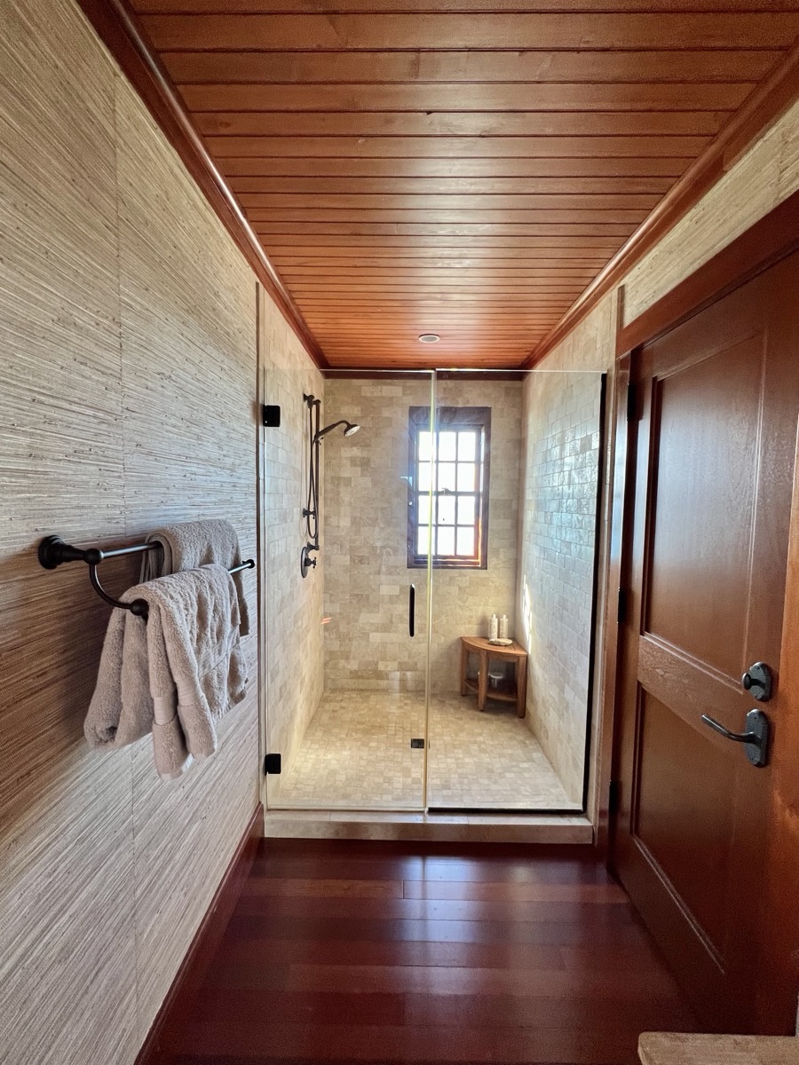 A glass walk-in shower with travertine tile, a rain head, teak bench and a garden window down a grasscloth hallway.