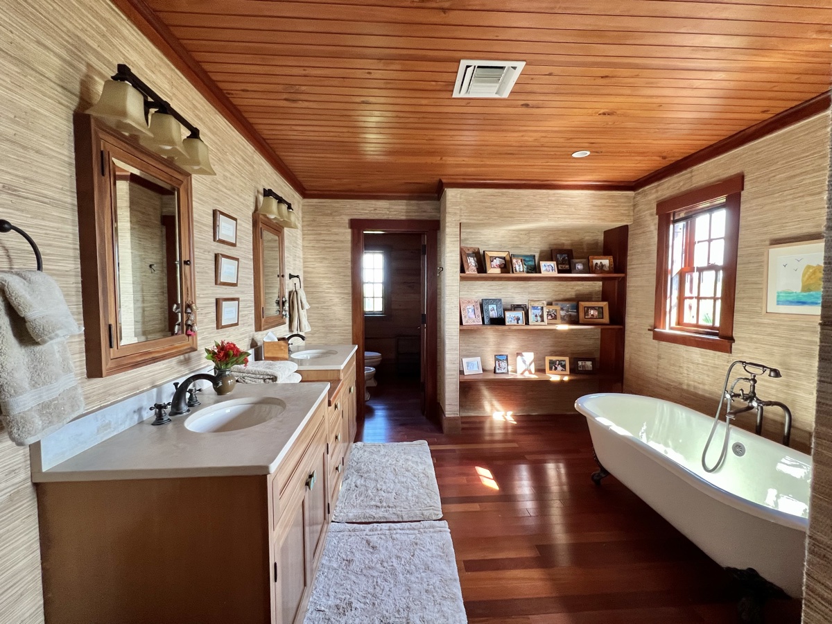 The master bath wide view with dual vanity, grasscloth walls, built-in shelves and clawfoot tub lit by natural light.