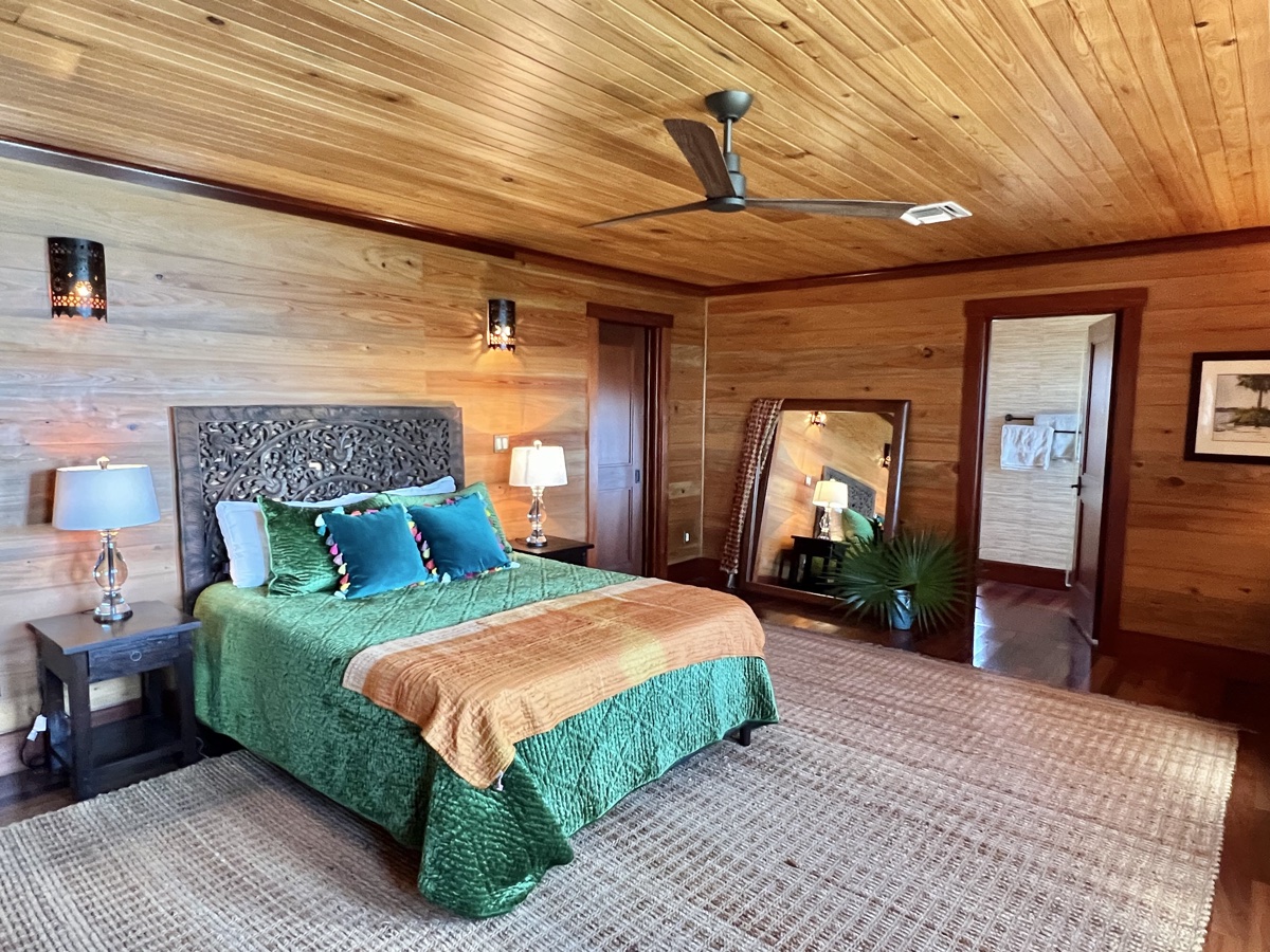 The primary bedroom with a carved headboard, green and gold linens, a floor mirror and warm cedar walls.