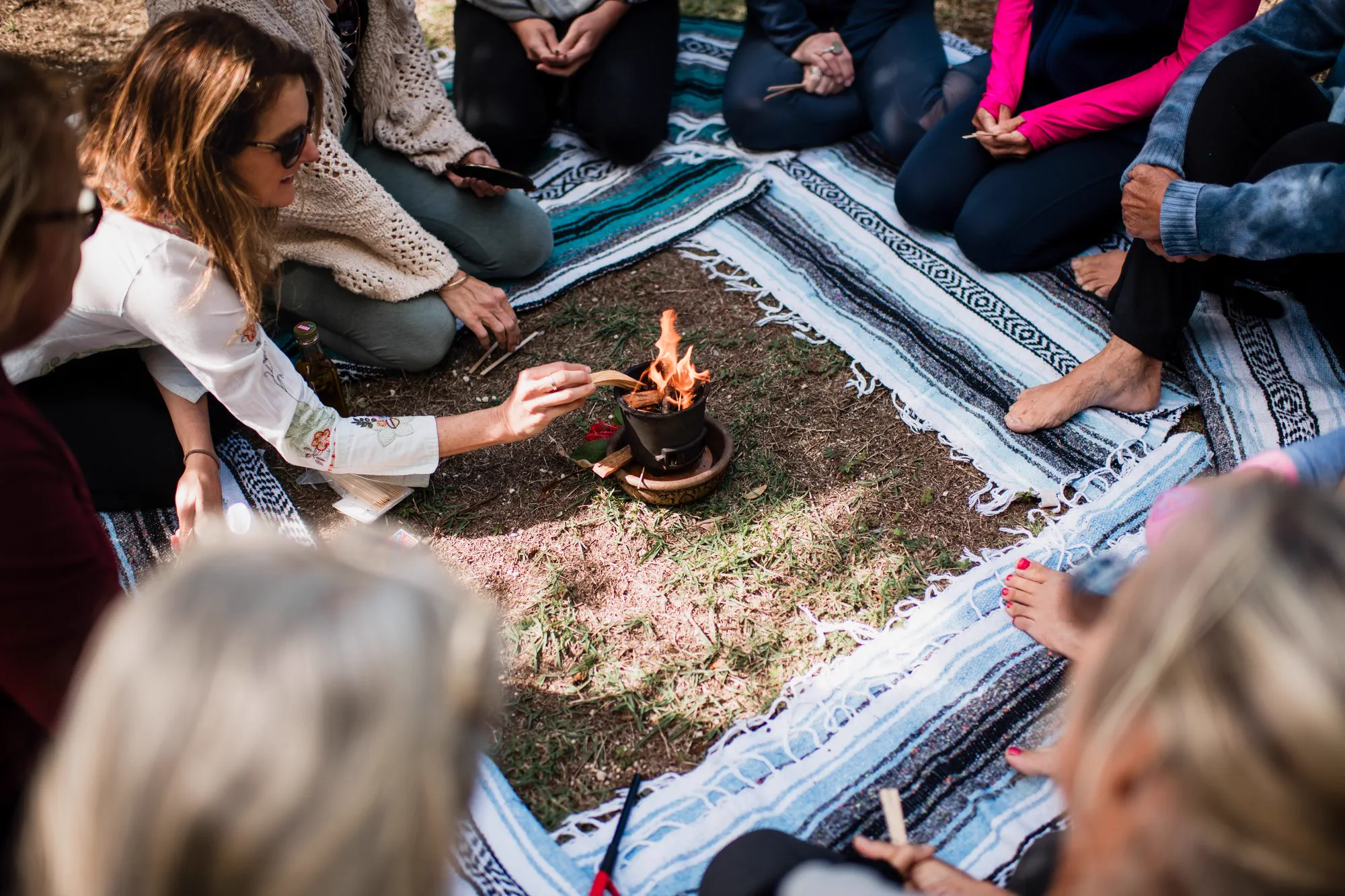 A small group seated in a circle on woven blankets around a ceremonial fire, one woman tending the flame.